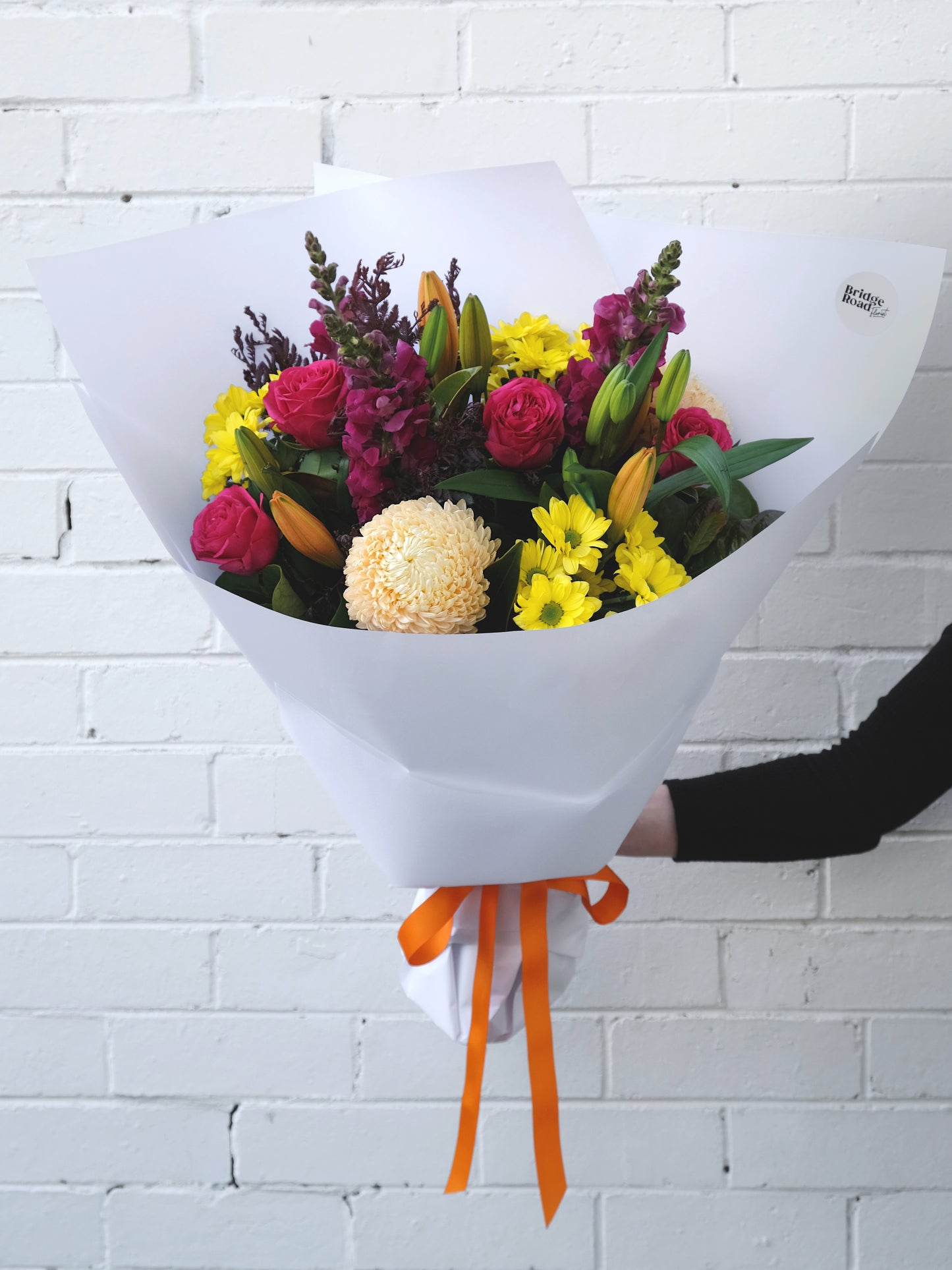 A person holding a bouquet of colorful flowers wrapped in white paper against a brick wall.