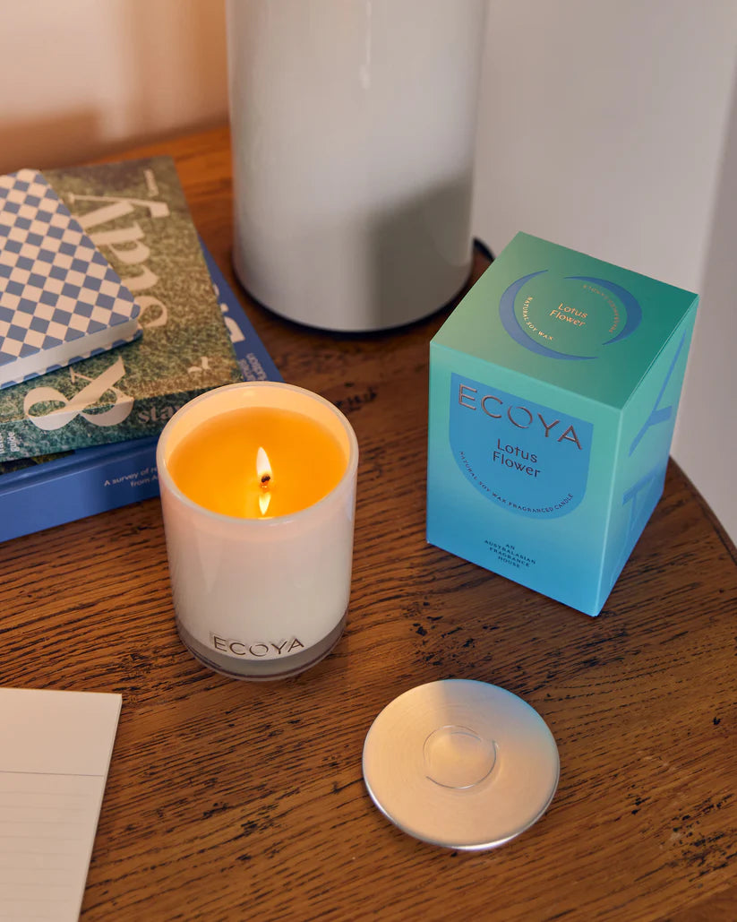 A lit candle on a table beside its packaging with books and a vase in the background.