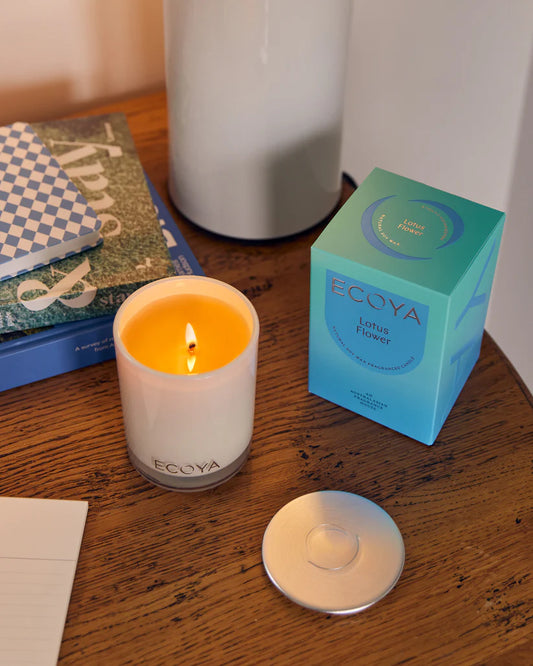 A lit candle on a table beside its packaging with books and a vase in the background.