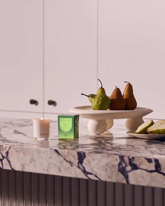 A lit candle, a box, and a fruit stand with pears on a marble countertop.
