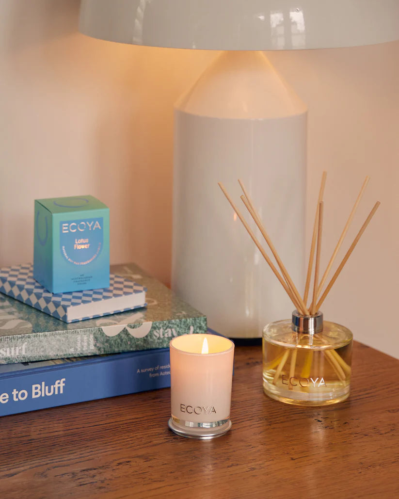 A lit candle, diffuser, and boxed product on a table with a lamp in the background.