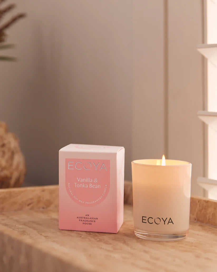 A lit candle next to its ECOYA Vanilla & Tonka Bean packaging on a wooden surface.