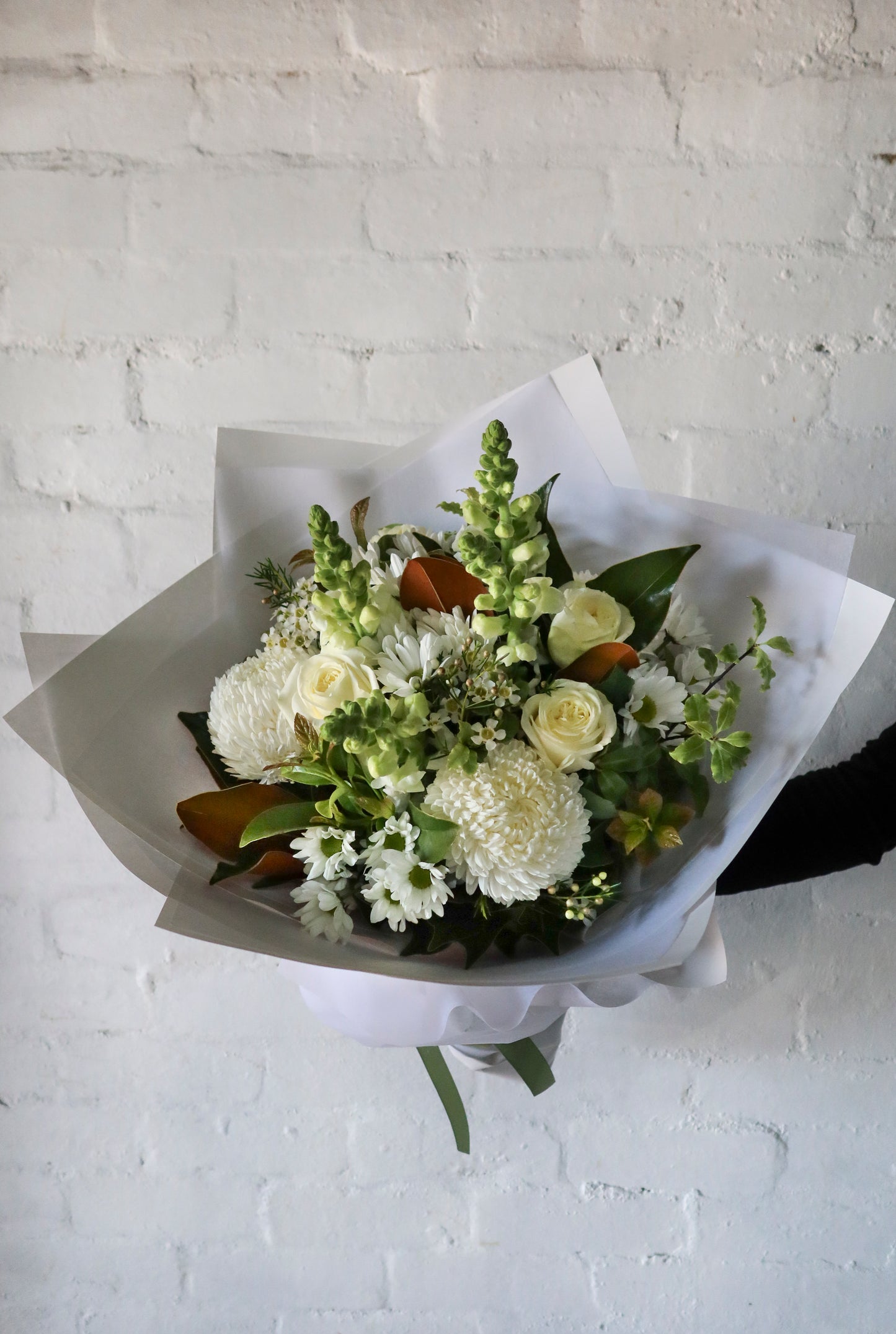 A bouquet of white flowers wrapped in paper against a white brick wall.