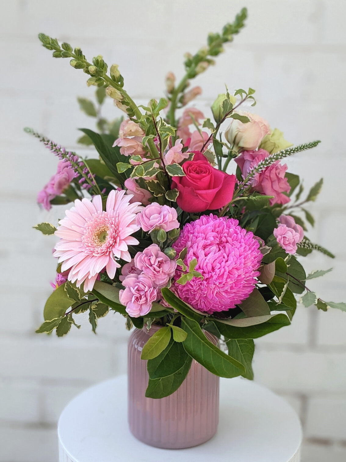 Pink floral arrangements against a white brick wall, one in a glass vase, the other in a purple pot.