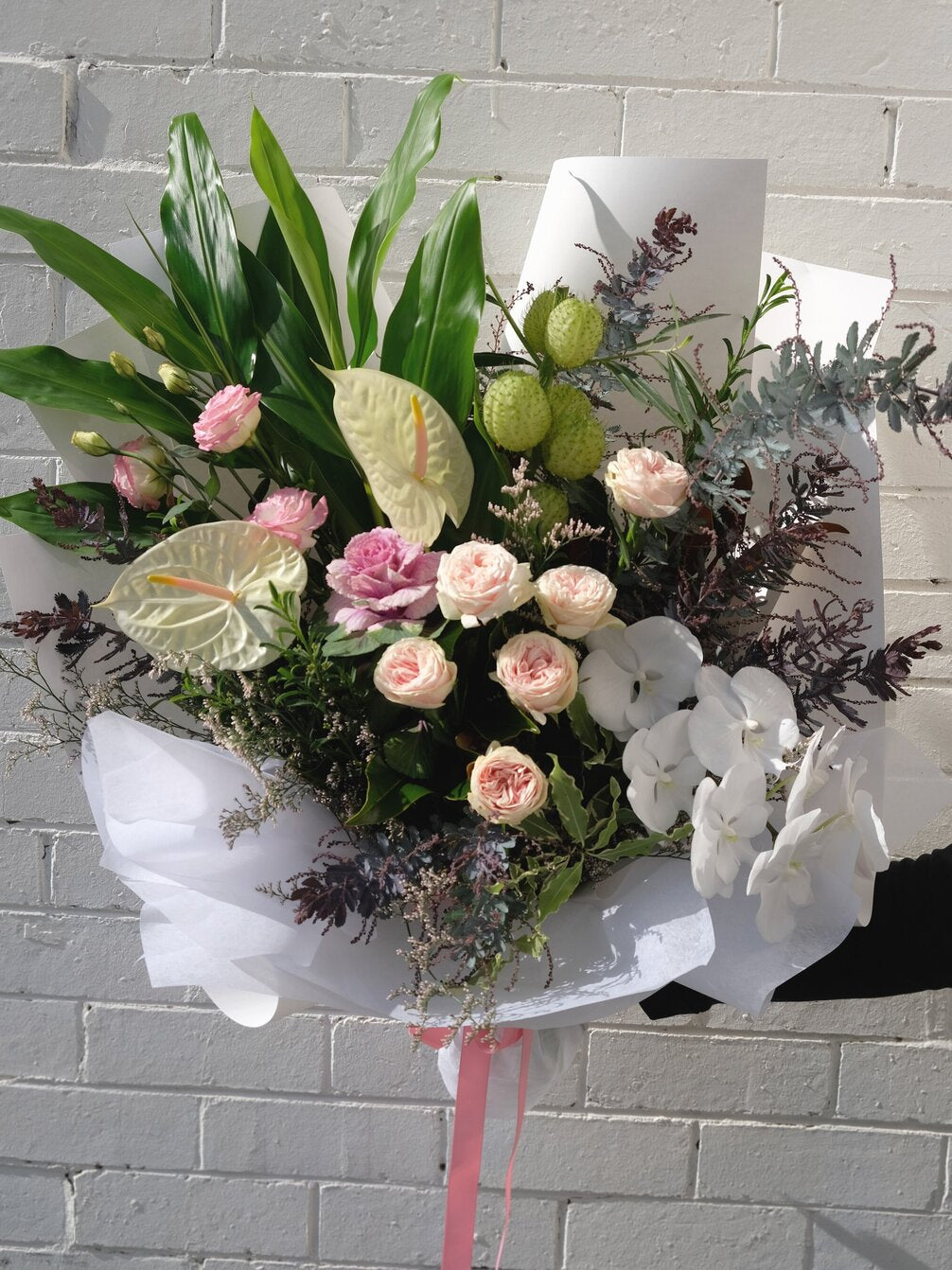 A bouquet of pink roses, white orchids, and assorted greenery against a brick wall.