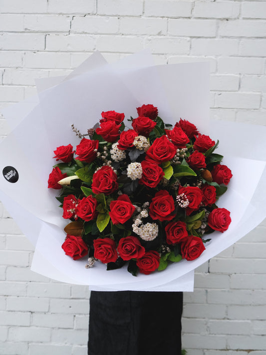A bouquet of red roses wrapped in white paper held in front of a white brick wall.