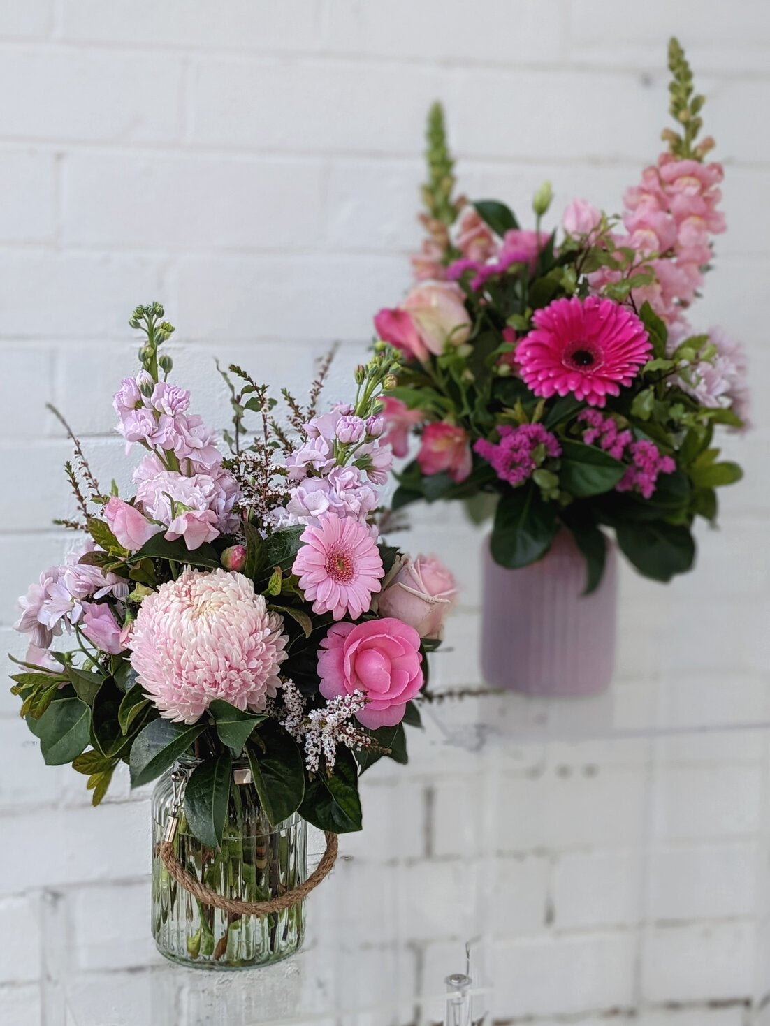 Two pink floral arrangements against a white brick wall, one in a glass vase, the other in a purple pot.