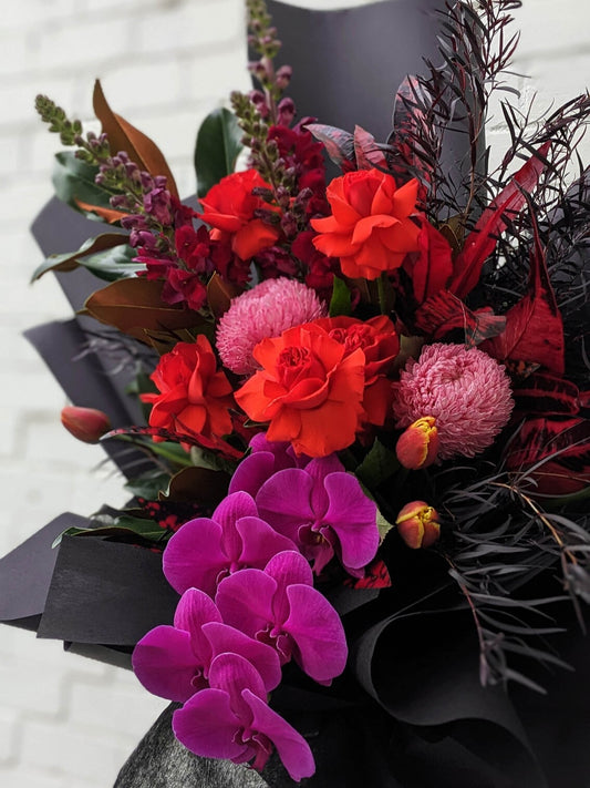 A vibrant bouquet featuring deep red roses, pink chrysanthemums, and striking purple orchids, surrounded by dark green foliage and elegant black wrapping.