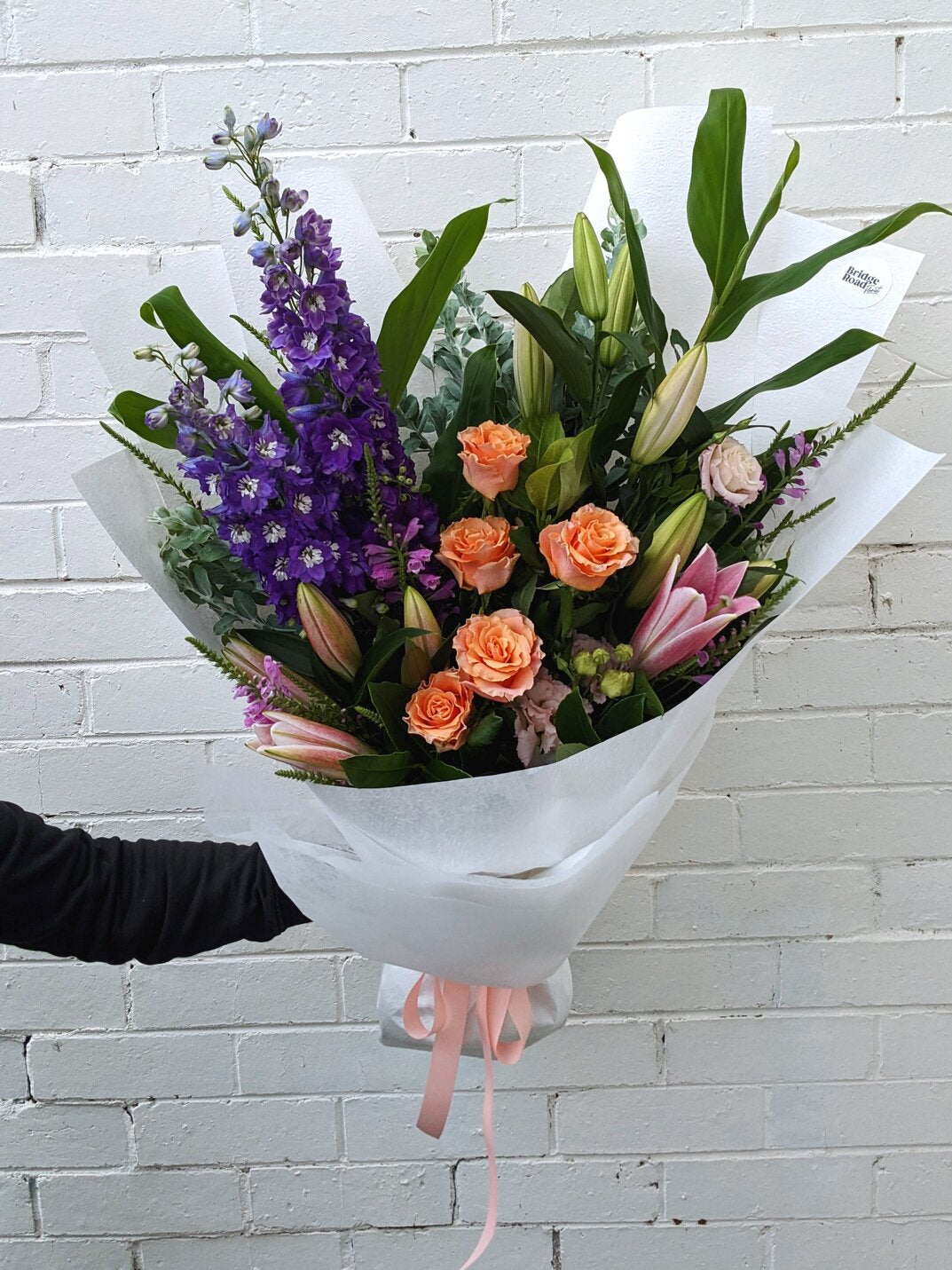 A vibrant bouquet of purple, pink, and orange flowers wrapped in white paper against a brick wall.