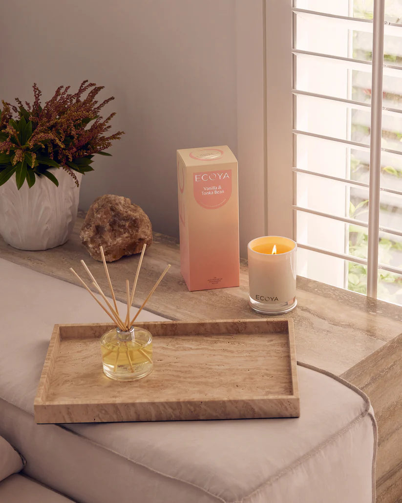 A reed diffuser on a wooden tray, a lit candle, and a box labeled "ECOYA" by a sunlit window.