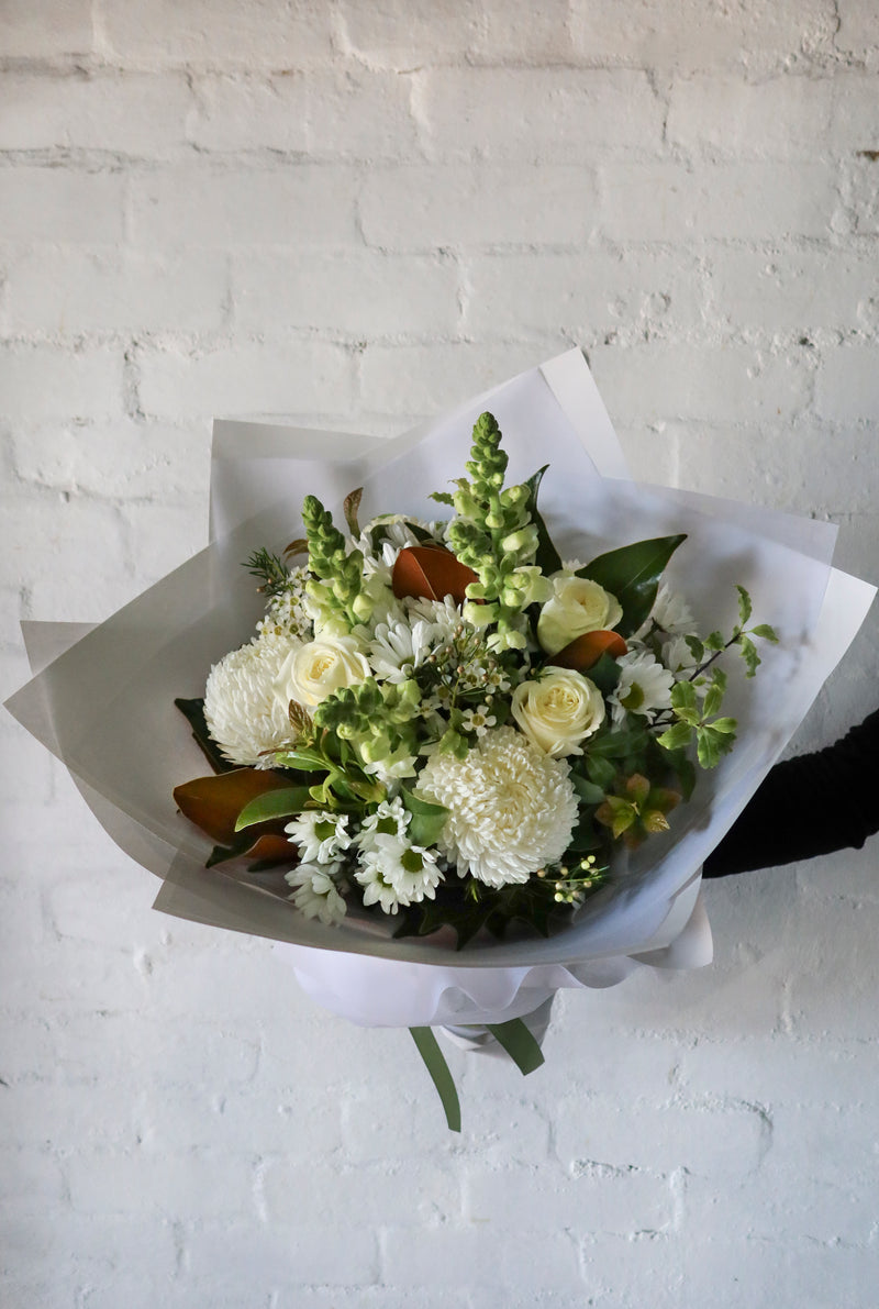 A bouquet of white flowers wrapped in paper against a white brick wall.