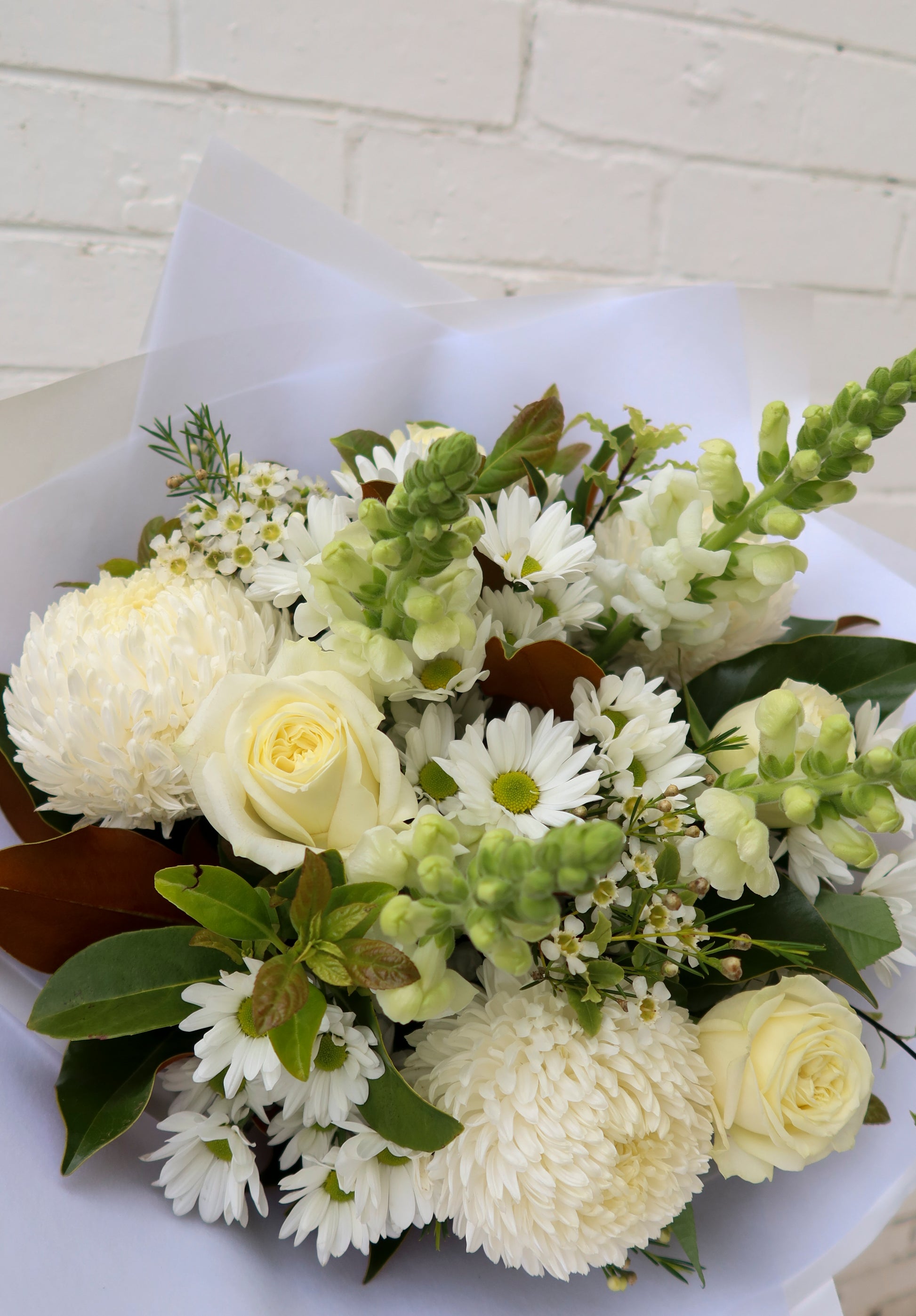 A bouquet of white flowers with green leaves wrapped in white paper against a brick wall.