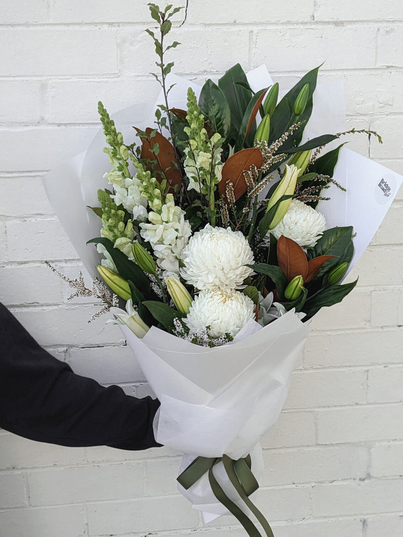 A person holding a bouquet of white flowers wrapped in white paper against a brick wall.