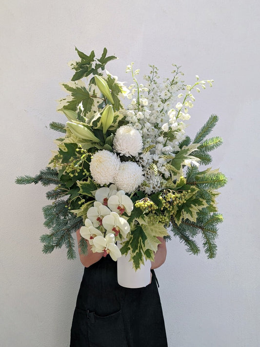 Person holding a large bouquet with white flowers and green leaves against a white wall.